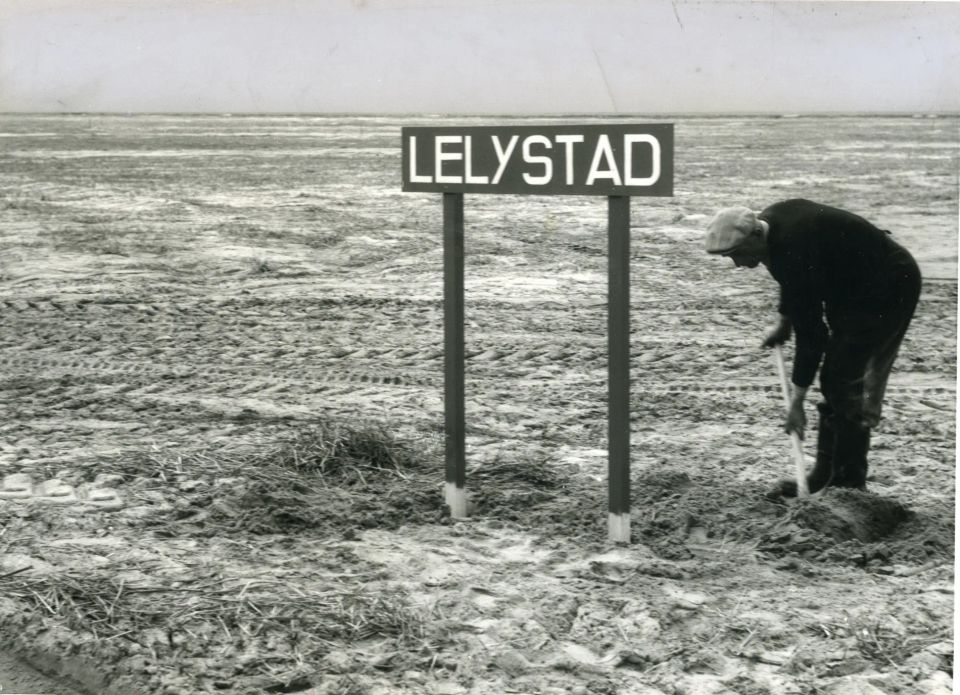 Een man zet een schep in de grond bij een bord van Lelystad
