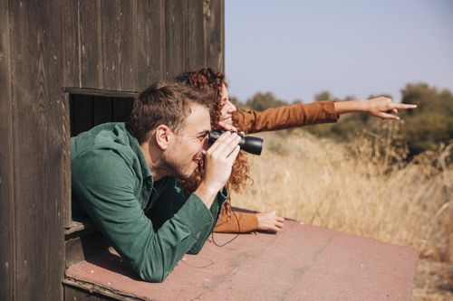 Een man en een vrouw zitten in een vogelspothut. De man heeft een verrekijker en de vrouw wijst naar iets in de verte.