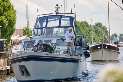 Er vaart een wit jacht met een oudere man en een vrouw erop in de doorgang van de brug van Delfstrahuizen, er wordt bruggeld betaald doormiddel van een hengel met een klomp. Op de achtergrond is een zeilschip te zien.