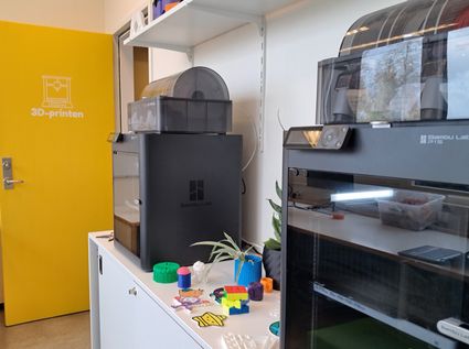 Een foto met in de voorgrond rechts twee 3D-printers. Schuin achter de printers is een gele deur die open staat, op de deur is een icoon van een 3D printer te zien met daaronder de text 3D-printen.