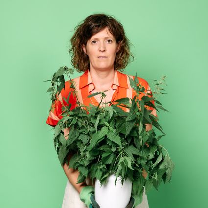 Foto van Janneke met een oranje bloesje aan tegen een groene achtergrond. Ze houdt een witte vaas met een grote bos brandnetels vast.