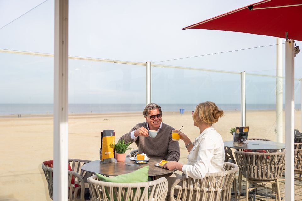 Echtpaar drinkt koffie met appeltaart op het terras van Strandpaviljoen Het Strand in Katwijk aan Zee, met uitzicht op zee.