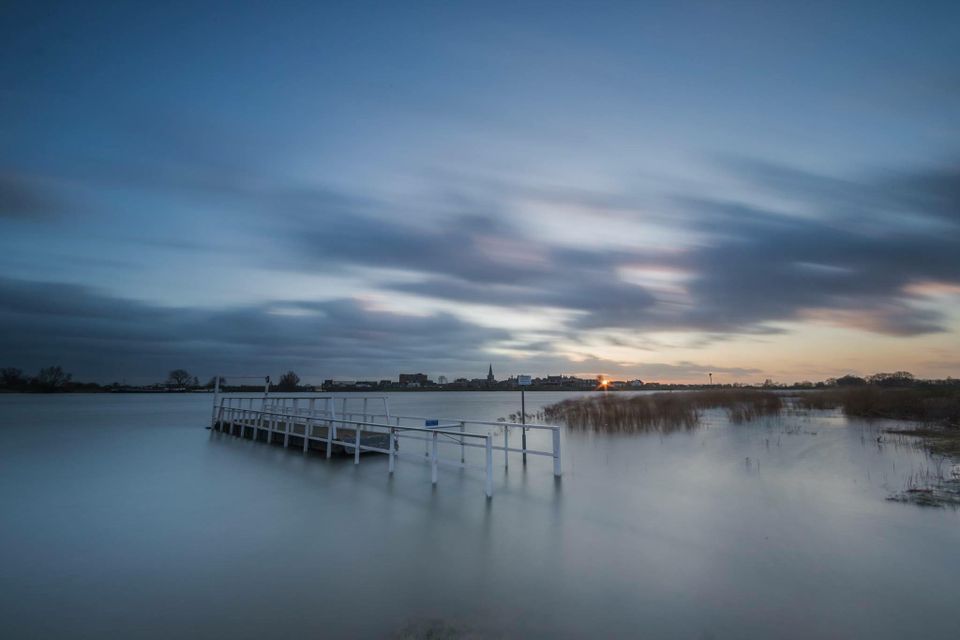 ChatGPT zei:

Natuurgebied Salmsteke in Lopik bij zonsondergang, met een witte steiger in het hoge water van de Lek, riet en overstroomde uiterwaarden, en in de verte de dorpssilhouetten met kerktoren onder dramatische wolken.