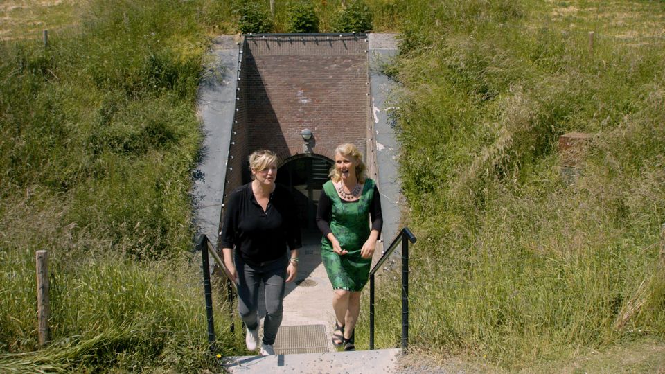 Twee vrouwen komen de bakstenen gang van een fort uitgelopen naar buiten. Achter de vrouwen zie je hoe het fort bekleed is met aarde en overgroeid met gras.