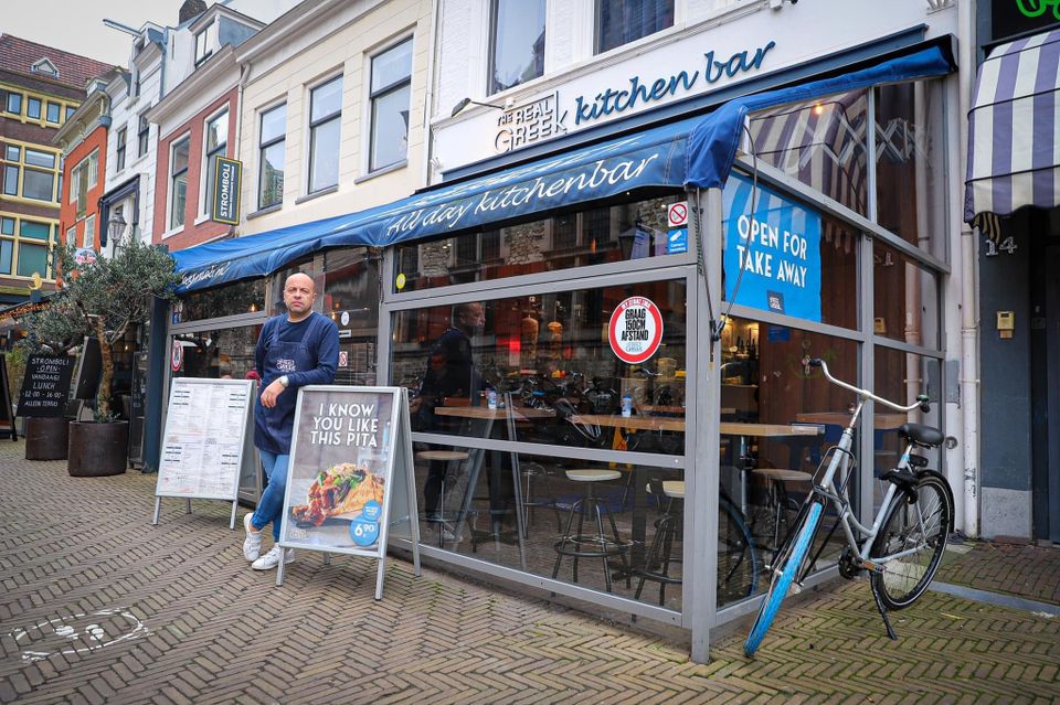 Vooraanzicht van het restaurant The Real Greek met de eigenaar ervoor