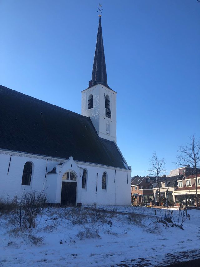 Het witte kerkje in Noordwijkerhout. Het is winter, de toren van de kerk steekt sterk af tegen de blauwe lucht. Er ligt sneeuw op de grond in de omgeving.