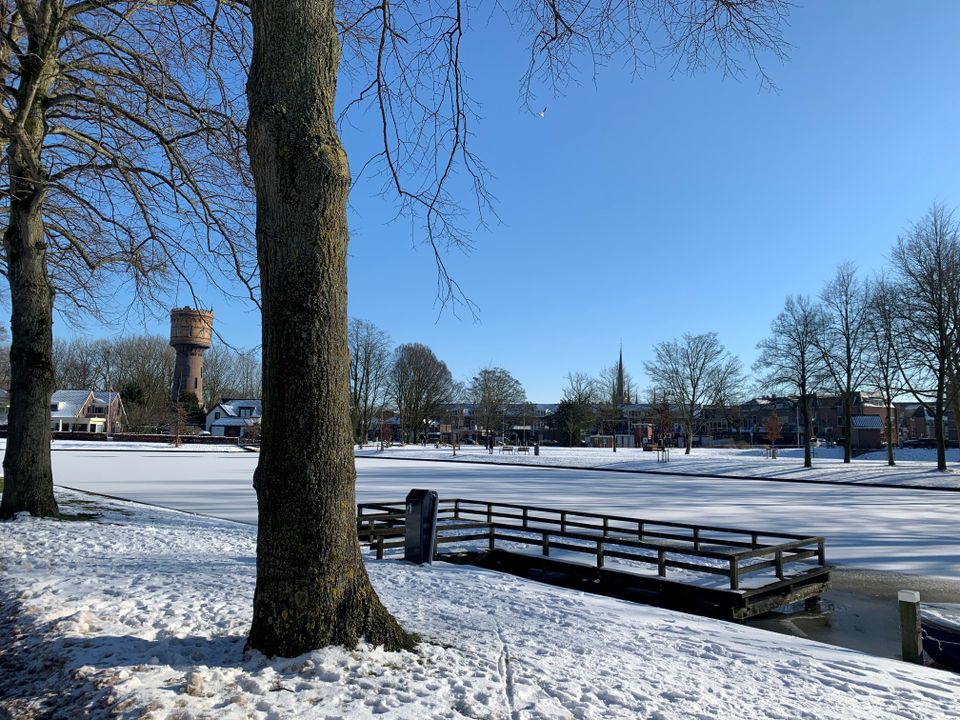 Een heldere winterochtend in Woerden, waar de singel volledig bevroren ligt onder een strakblauwe lucht. Tussen de kale bomen kijk je uit op de karakteristieke watertoren en de kerktoren in de verte. De combinatie van zonlicht, sneeuw en stilte geeft het tafereel een bijna magische, rustige sfeer.