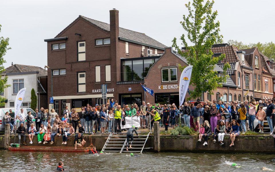 Sportevenement Triathlon Woerden, Woerden, Groene Hart, zwemonderdeel in de singel met triatleten in wetsuits die via een trap uit het water klimmen terwijl veel publiek langs de kade en bij winkels in het centrum toekijkt.