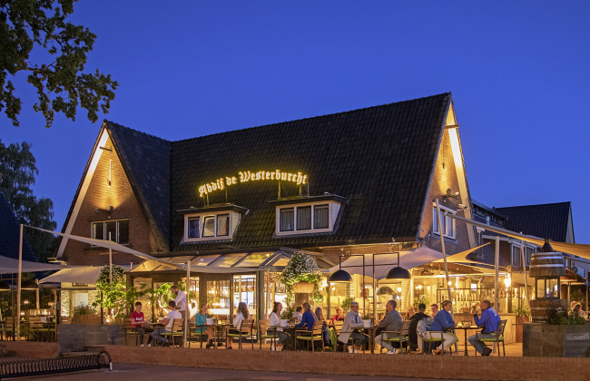 Mensen genieten in de avonduren van een hapje en een drankje  op het verlichte buitenterras van hotel en restaurant Abdij de Westerburcht.