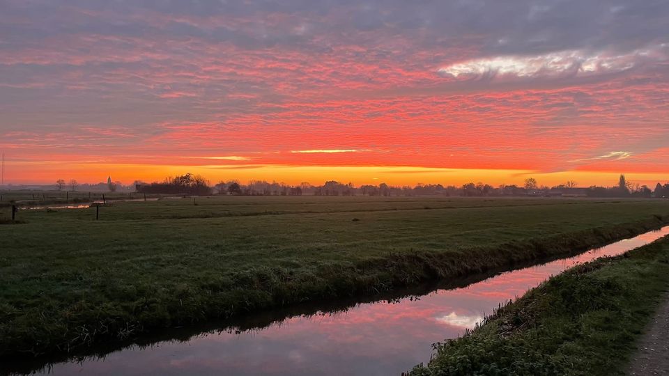 Polderlandschap, Lopik, Groene Hart, uitgestrekte weilanden en sloten tijdens een kleurrijke zonsondergang met rood-oranje lucht en silhouetten van bomen en bebouwing aan de horizon.