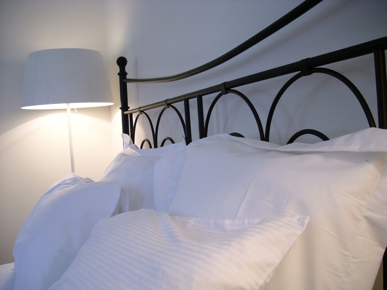 Detailfoto van een met wit beddengoed opgemaakt bed, met siersmeedwerk hoofdeinde en een witte, brandende schemerlamp.