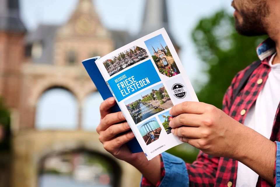 Reisgids Friese Elfsteden in iemands handen met op de achtergrond de Waterpoort in Sneek.