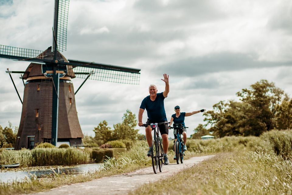 Polderlandschap met windmolen bij Alphen aan den Rijn, Groene Hart, stel senioren fietst over smal dijkpad langs watergang en rietkragen tijdens zomerse dag met bewolkte lucht.