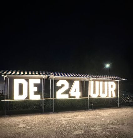 Verlicht logo van de 24 Uur van Woerden met grote lichtgevende letters ‘DE 24 UUR’, opgesteld langs een weg in het donker, onderdeel van het evenement 24 Uur van Woerden.