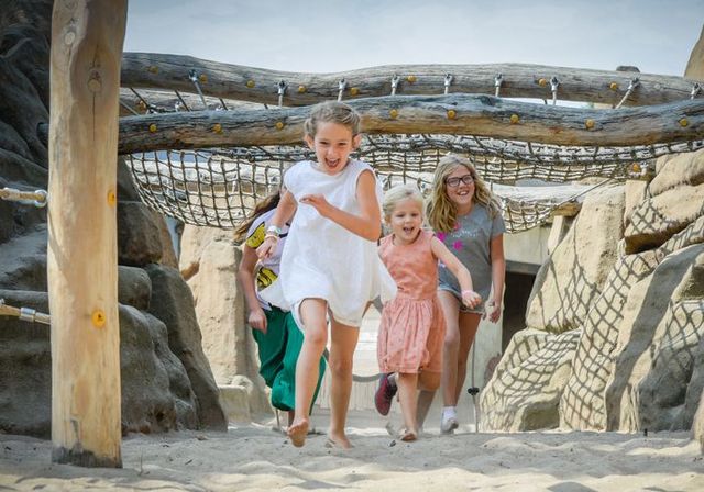 Kinderen rennen lachend door de avontuurlijke speeltuin van Vogelpark Avifauna in Alphen aan den Rijn, omgeven door rotsen, zand en touwbruggen.