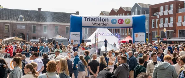 Drukke finishlocatie van de Triathlon Woerden op het Kerkplein met sporters die onder de blauwe finishboog doorlopen en omringd door een groot publiek en festiviteiten.
