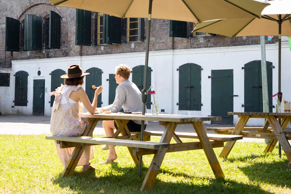 Een man en een vrouw zitten op een picknickbank in het zonnetje, voor een fort.