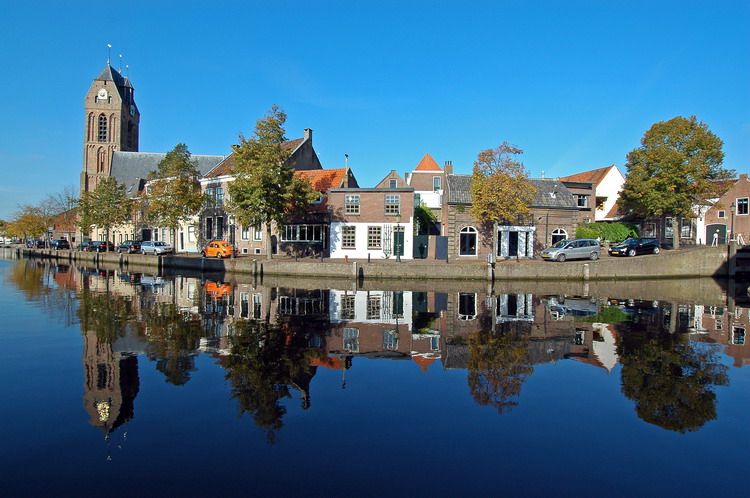 Historische gevels en de Grote Kerk van Oudewater langs een stille gracht, met spiegeling in het water en een heldere blauwe lucht.