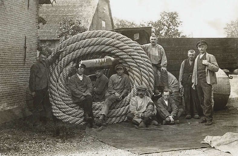 Historische zwart-witfoto van touwslagers bij het Touwmuseum in Oudewater, met een grote opgerolde tros touw en arbeiders die poseren op het erf van de touwslagerij.