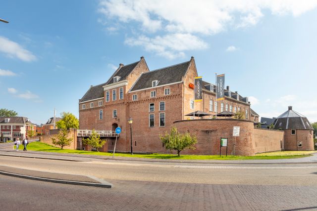 Zijaanzicht van Kasteel Woerden met zijn robuuste bakstenen muren, vlaggen met de tekst Woerden en wandelende bezoekers op een zonnige dag.