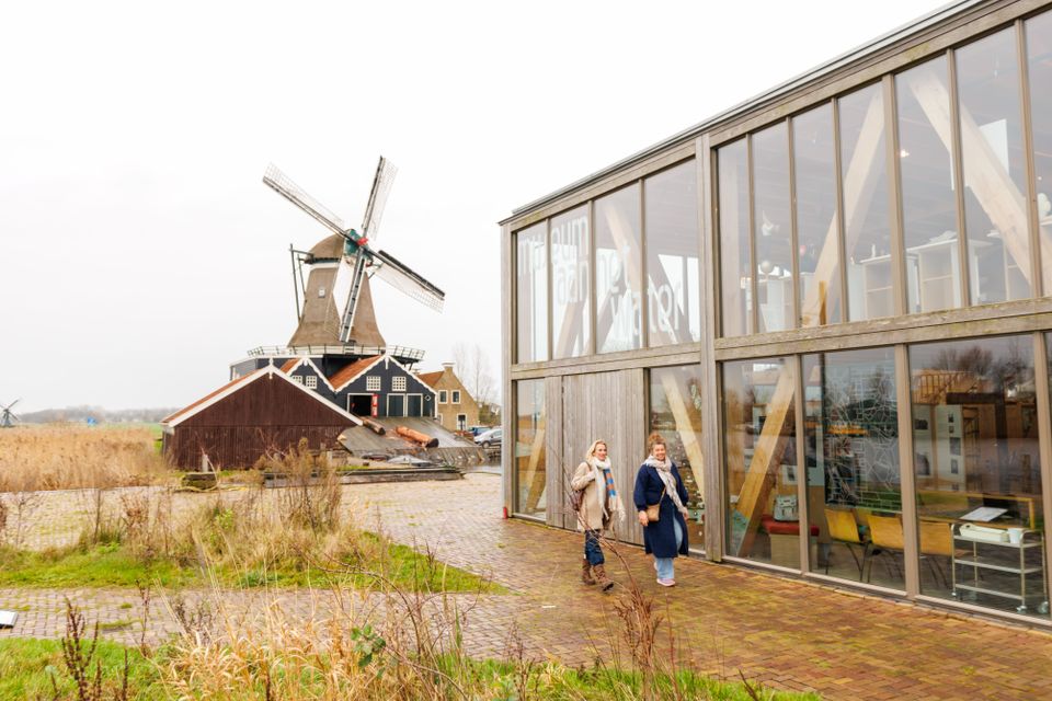 Twee mensen lopen buiten op het terrein bij Museum Houtstad IJlst. Op de achtergrond zie je Houtzaagmolen de Rat in IJlst.