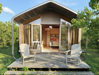 Klein duurzaam wikkelhuisje met open deuren, houten stoelen op het terras en uitzicht op een groene boomgaard bij de Proeftuin van Linschoten in de omgeving van Woerden.