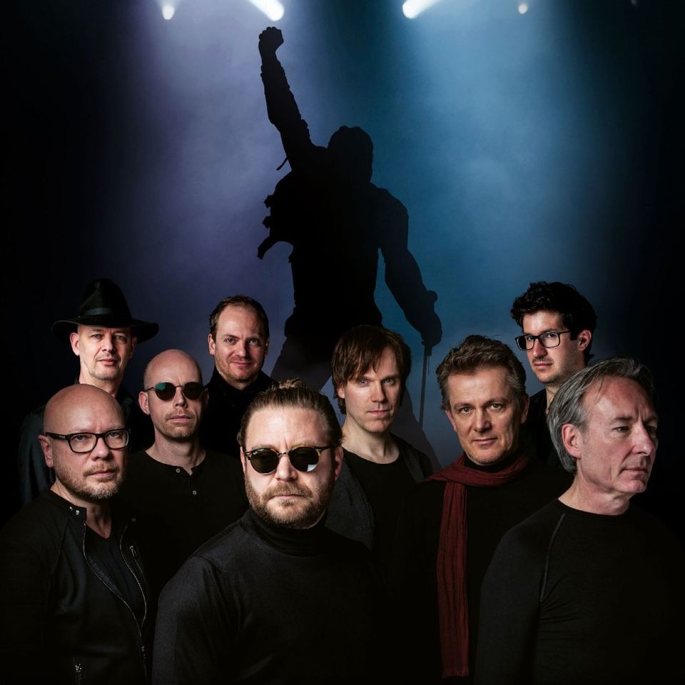 Donkere foto van de uit 9 mannen bestaande band in donkere kleding met op de achtergrond een silouet van een persoon die een optreden geeft.