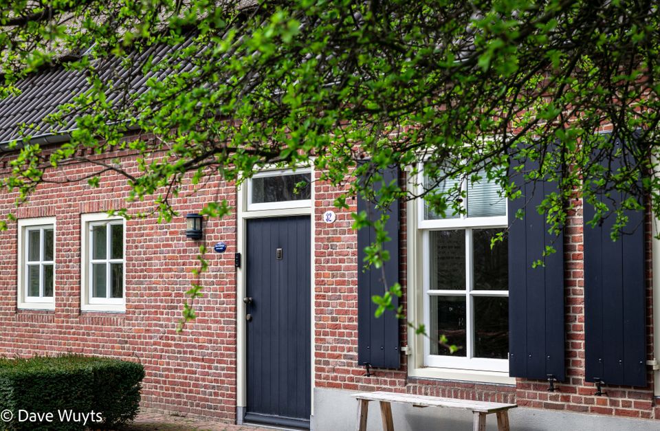 Gevel van een boerderij, donkere voordeur en luiken. Houten bankje onder het raam en de bladeren van een boom op de voorgrond.