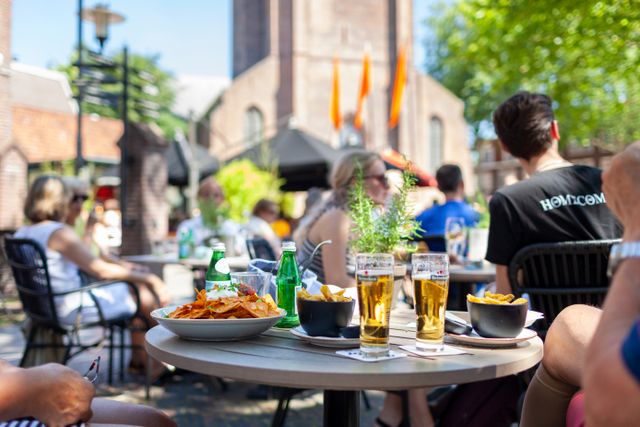 Gezellig terras met mensen die genieten van drankjes en hapjes op een zonnige dag voor de Petruskerk in Woerden.