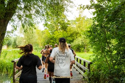 Natuurgebied in Woerden, Groene Hart, social run van de Offline Outside Social Club met hardlopers die over een houten brug door een groen park en langs het water rennen tijdens een gezamenlijke hardloopactiviteit.