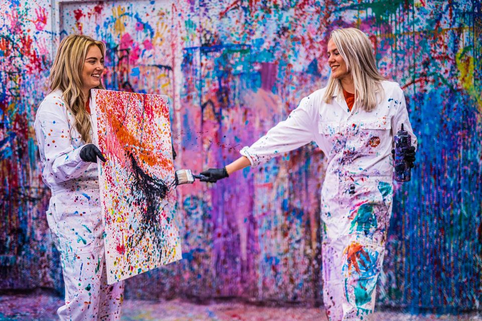 Twee vrouwen in witte overalls maken een kleurrijk schilderij door verf te spatten tijdens de creatieve activiteit Gooien met Verf bij Fabriek Woerden.