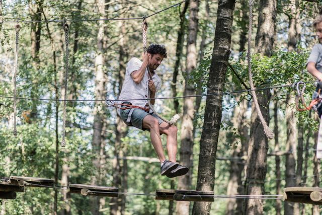 Een man slingert in het klimpark met een touw van plank naar plank.