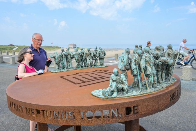 Twee wandelaars bekijken het kunstwerk Kalla's Toren, van kortenstaal en ander metaal. Een beeldengroep staat op een metalen tafel.