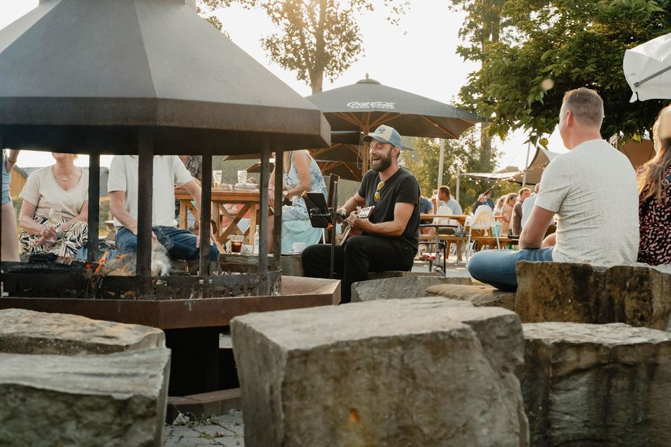 Man speelt gitaar en zingt bij een vuurkorf op een terras, terwijl bezoekers op banken en aan tafels luisteren en drinken. Informele buitensfeer met parasols en zomers licht, wat duidt op een kleinschalig live optreden.