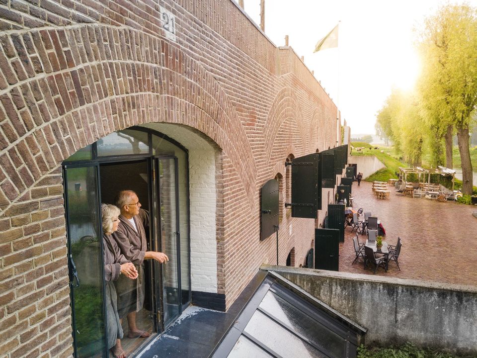 Een man en een vrouw staan in hun badjassen op een Frans balkonnetje, gemaakt in een oud raamkozijn van een bakstenen fort. Ze kijken uit over de binnenplaats.