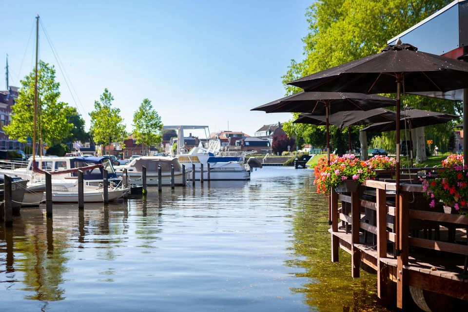 Zicht op de haven van Woerden met aangemeerde bootjes en een terras met bloembakken en zwarte parasols aan het water op een zonnige dag.