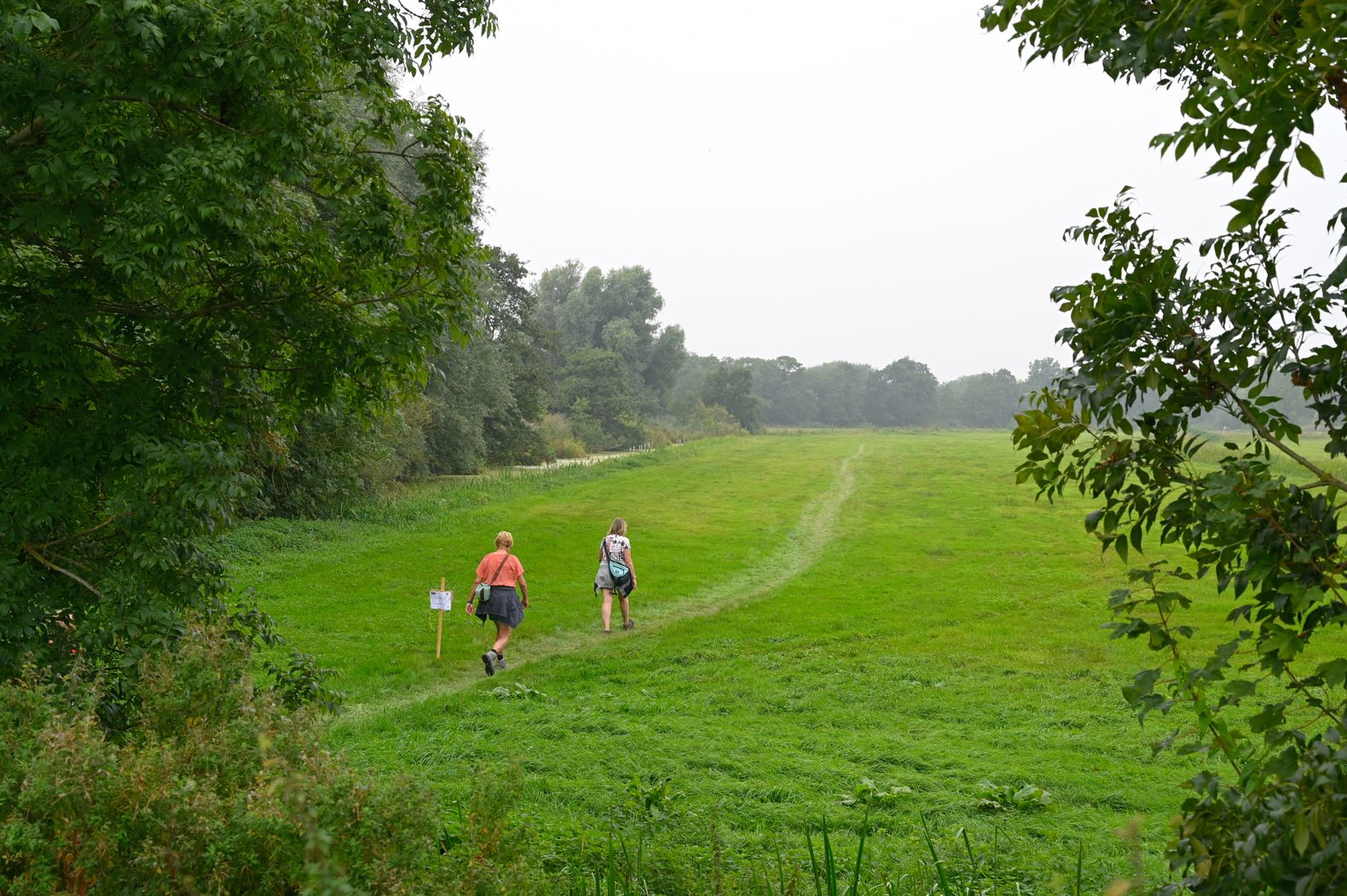Een man en een vrouw die over een wandelpad door het grasveld lopen wat omringt is met bomen en struiken.