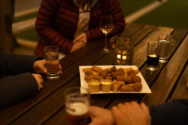 Gezellig borrelmoment op een houten terras met warme sfeerverlichting, waar een groep mensen geniet van een royale schaal bittergarnituur met sauzen en diverse drankjes