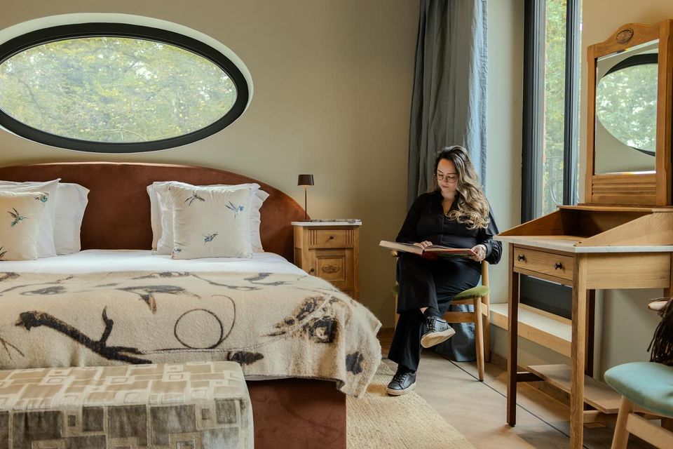 Een ruime, gezellige hotelkamer met liggend ovaal raam boven een groot bed en een dame die zit te lezen in de stoel naast het bed.