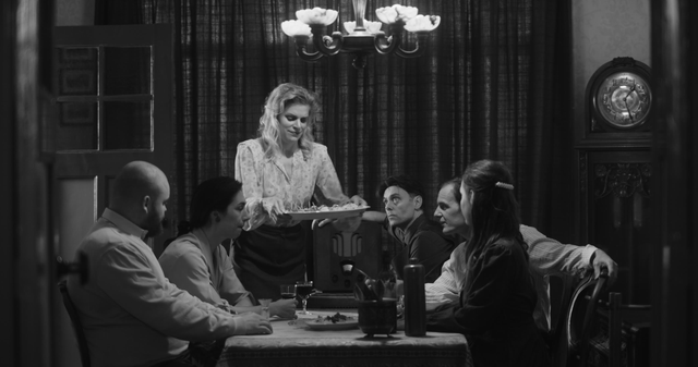 Still scene uit film Bezuidenhout in zwart wit. Vrouw zet schaal met voedsel op tafel, waaraan haar gezin zit.
