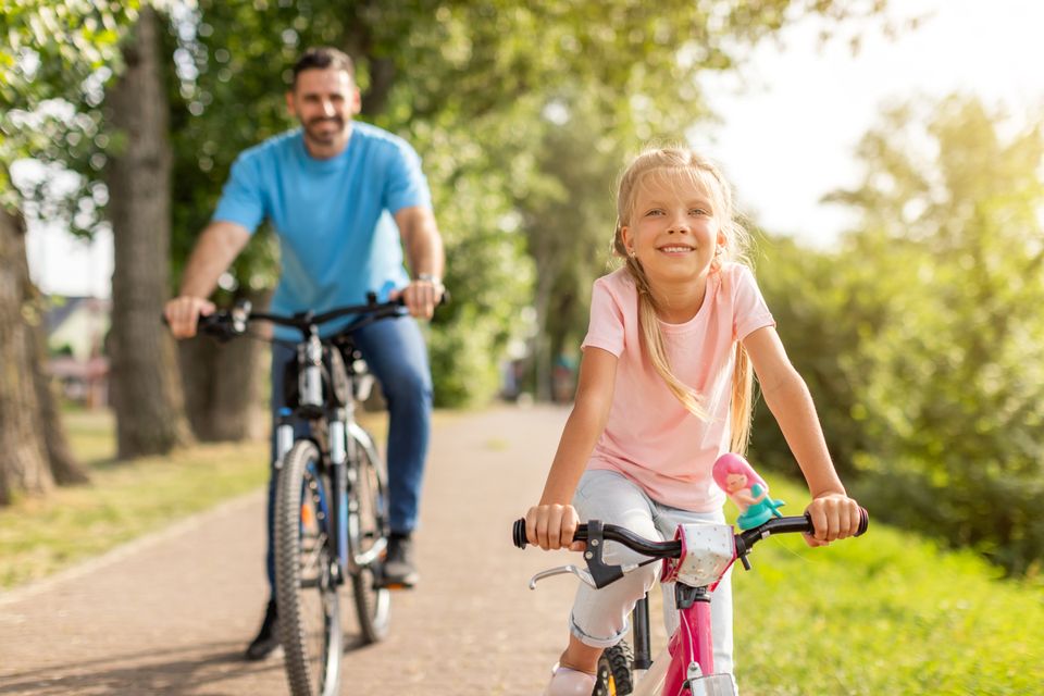 Meisje fietst voorop over een zonnig pad door het groen, terwijl een man op een fiets achter haar volgt; samen genieten ze van een ontspannen fietstocht in de natuur.