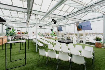 Binnenaanzicht van KASXL, een moderne en lichte vergader- en evenementenlocatie van KAS Meeting & Eventlocatie. De ruimte is ingericht met rijen witte stoelen gericht op een podium met schermen waarop “AI en events” staat. De locatie heeft een glazen kasconstructie, kunstgrasvloer, hoge tafels, veel daglicht en is omgeven door planten en stadsuitzicht.