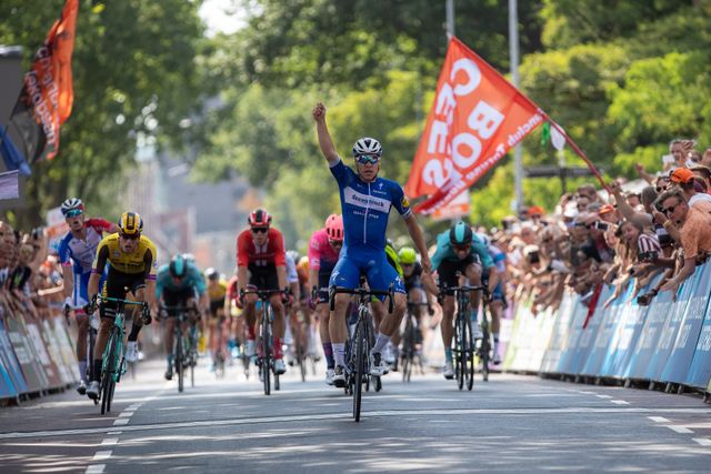 Een wielrenner komt met gebalde vuist over de finish terwijl hij het peloton achter zich laat.