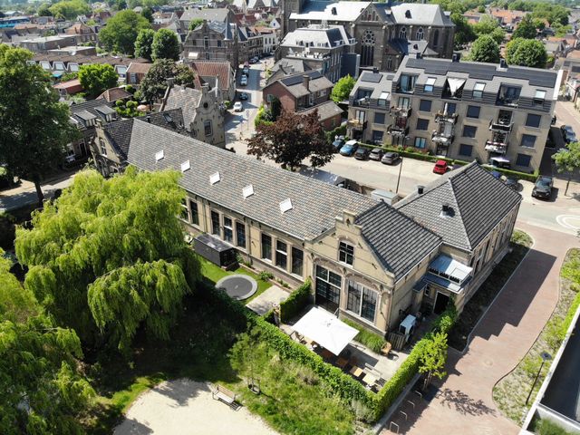 Culturele ontmoetingsplek Muziekhuis in Oudewater, Groene Hart, historisch gebouw met grote ramen en terras gezien vanuit de lucht, omringd door groen park, woonhuizen en de binnenstad met kerk op de achtergrond.