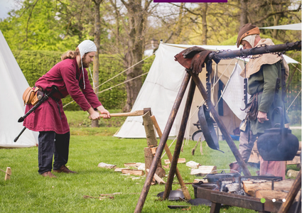 Twee personen in middeleeuwse kleding bij een kampement met tenten. Eén hakt hout met een bijl op een blok, de ander staat bij een vuur met kookgerei. Op de grond liggen houtblokken in een groene buitenomgeving.