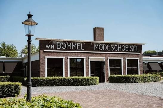 Foto van een plat stenen gebouw met grote ramen en de letters Van Bommel op de gevel. Omringd met groene haagjes.