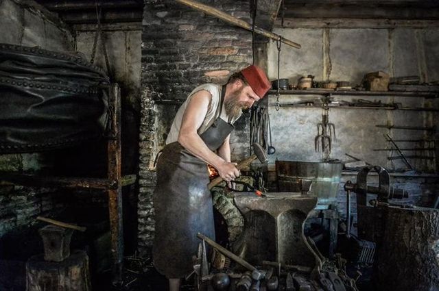 Middeleeuwse smid bewerkt gloeiend ijzer op een aambeeld in een historische smederij in Archeon, omringd door traditionele werktuigen en gereedschap.