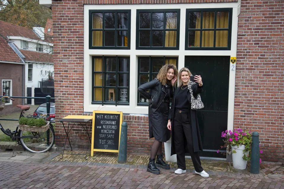 Twee lachende vrouwen die een selfie maken voor een klein, historisch pand met een bord dat "Het kleinste restaurant van Nederland" aangeeft