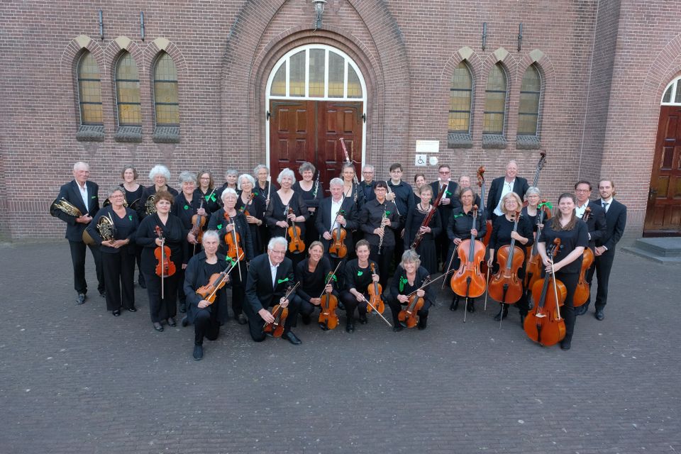 Culturele muziekuitvoering Het Groene Hart Orkest in Woerden, Groene Hart, groepsfoto van muzikanten met violen, cello’s en blaasinstrumenten voor bakstenen kerkgebouw tijdens concert of repetitie.