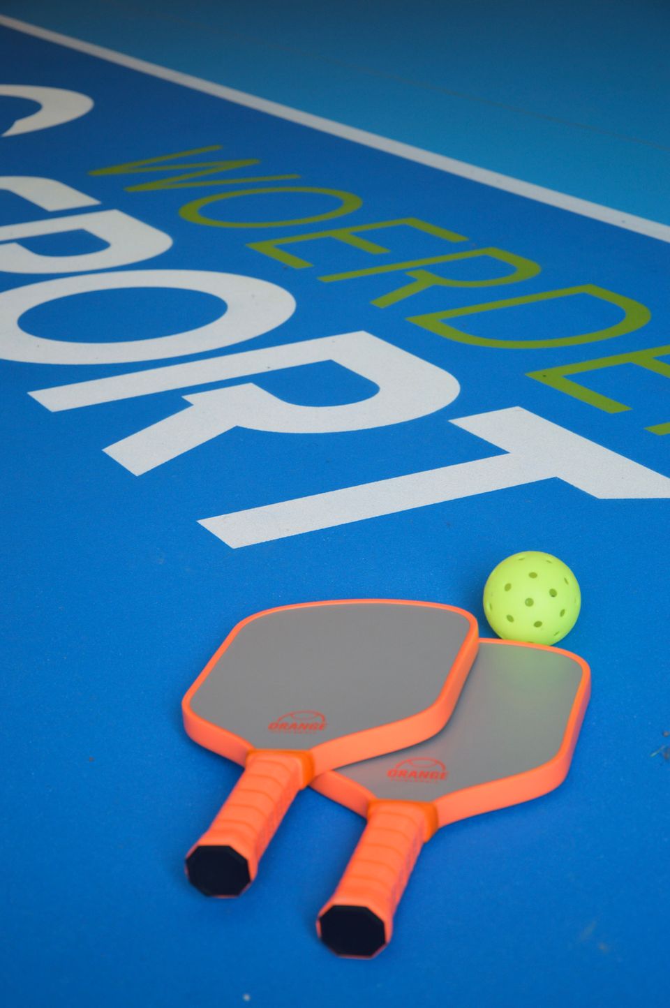 Pickleballbaan bij Zwembad H2O in Harmelen, Groene Hart, met twee pickleballpaddles en een gele geperforeerde bal op de blauwe sportvloer.